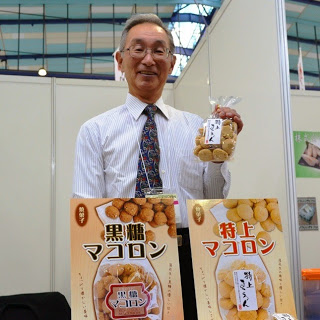 白いシャツにネクタイ姿の男性が、「黒糖マコロン」や「特上マコロン」と書かれたパッケージと共に袋入りのマコロンを持って紹介している写真