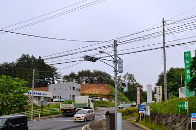 坂道のある交差点付近に車が行き交い、奥に開発途中の茶色い土の斜面と白とグレー色の外壁の建物が見える写真