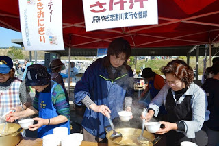 赤いテントの下で、女性たちが湯気の立つ大鍋から汁物をよそい、白い器に入れて配っている写真