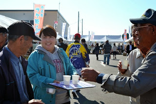 紙コップに入った飲み物を持った女性が、来場者の男性たちに笑顔でふるまっている写真