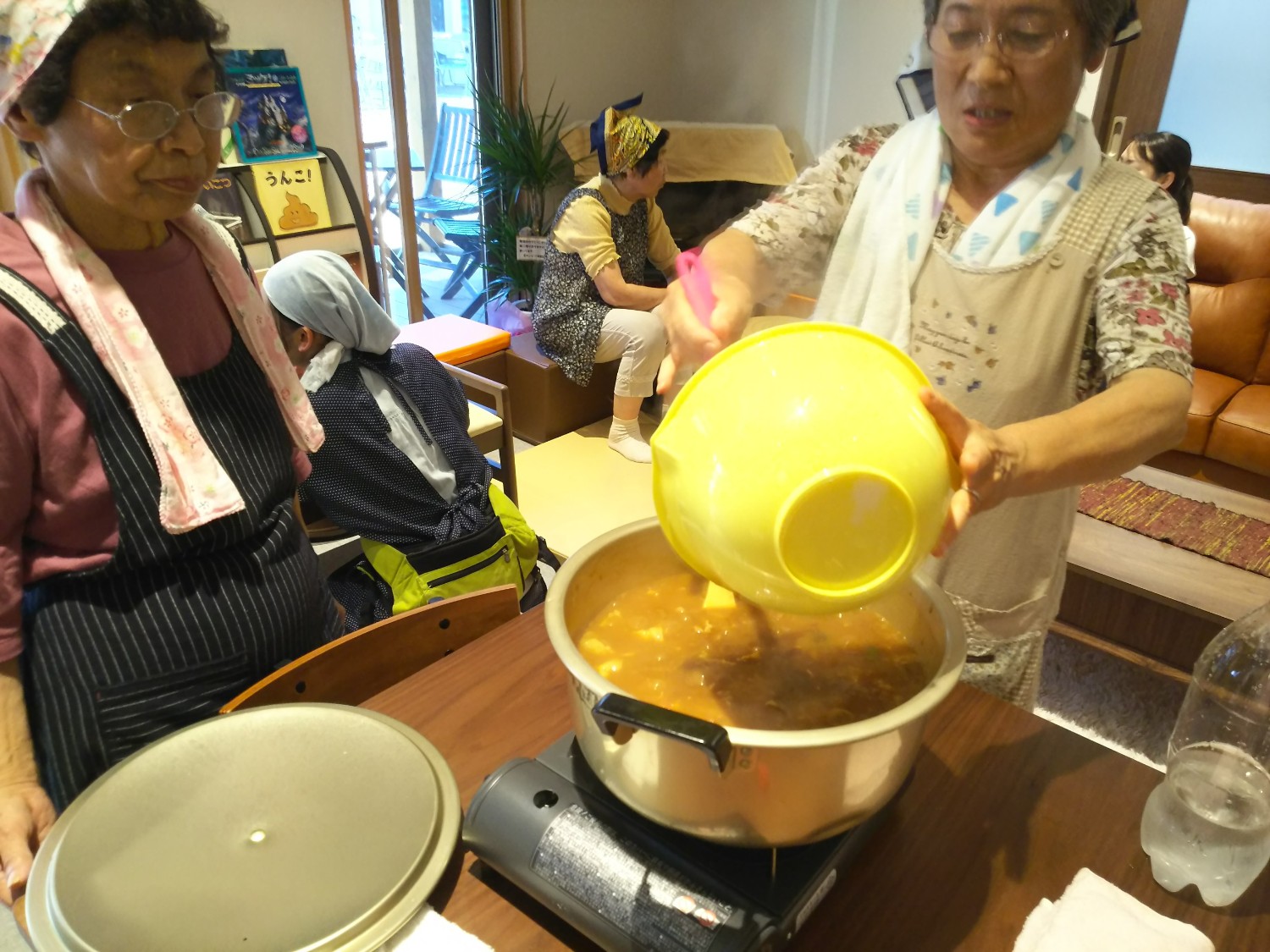 室内のテーブルで、三角巾とエプロンを着けた年配の女性が、大きな黄色のボウルから具材を鍋に入れてカレーを作っている写真