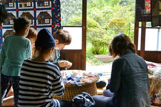 窓の外には緑の庭が広がっている古民家の室内で、女性が手作業をしている様子を小さな子供がのぞき込んで見ている写真