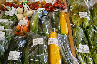 にんじん、ズッキーニ、トマト、にんにく、葉物野菜などがひとつずつ透明な袋に詰められ、値札シールが貼られて木の台に並べられている写真