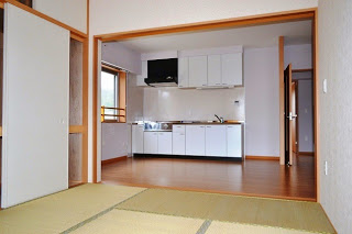 左側に窓、右奥に出入口の扉があり、天井下に収納スペースが兼ね備えられた白いシステムキッチンと、フローリングのダイニングが広がっている様子を畳の部屋から見た写真