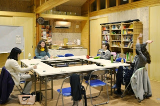 木の温もりを感じる部屋で4人の女性が机を囲んで座り、両手を挙げたり笑ったりしながら自由な雰囲気で会話を楽しんでいる英会話教室の写真