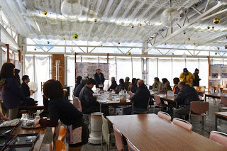 室内の前方で挨拶をしている人がたち、その前に参加者の人たちが机に座って説明を聞いている写真