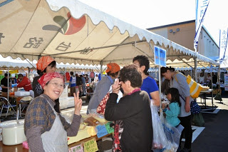 2人のスタッフが買い物客と楽しげに会話をしている傍で、頭にバンダナを巻いた女性店員がカメラに向かって笑顔でピースサインをしている写真