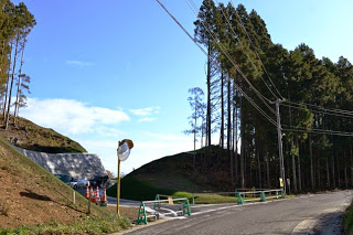 木が立ち並ぶ中に舗装された道と工事用バリケードが置かれている、山を切り開いて道路を通している造成地の入口の写真