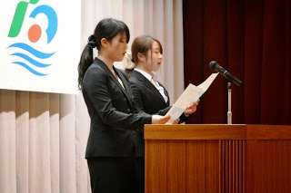 スーツ姿の二人の女性が並んでマイクの前で両手に紙を持ち町民憲章を唱和している写真