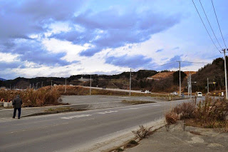 道路と歩道の境目に人が立ち、奥には造成中の高台や電柱が立ち並ぶ風景が広がっている写真