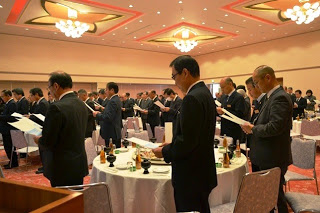 シャンデリアのある宴会場で、黒いスーツ姿の男性たちが起立し、同じ紙を手にして町民憲章を唱和している写真