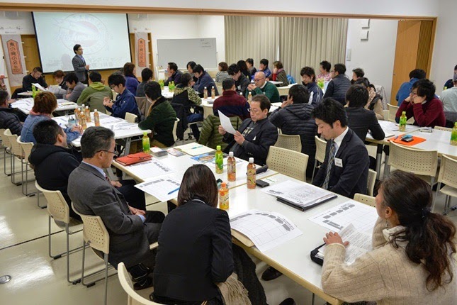 会議室のような部屋で、複数のテーブルに分かれて座った多くの参加者が資料を見ながら、スクリーンの前に立つ男性の話を真剣に話を聞いている写真