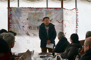 手づくりの幕の前で男性が紙を持ちながら挨拶をしている写真