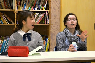 本棚の前に座った左側の女性が口をあけて先生が座っている右側を向き、右側に座っている先生がぬいぐるみを手にして身振りを交えて説明している様子の写真