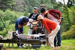 屋外のベンチの上に置かれたドローンの操作機材を囲みながら、数人の大人が画面を真剣にのぞき込んでいる写真