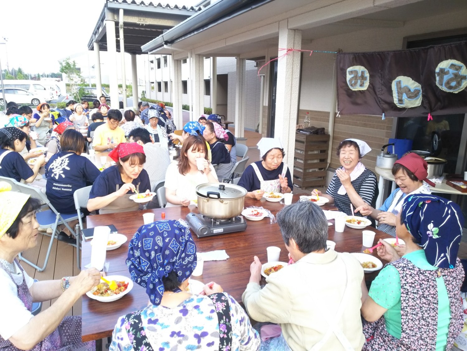 屋外のテーブルを囲んで多くの参加者たちが完成したカレーライスを食べながら楽しそうに会話している写真