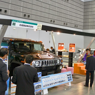 黒い大型車両と共に企業ブースが設けられ、数人の男性が説明を受けながら資料を見る様子を写したビジネス展示ブースの写真