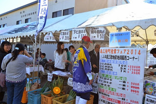 白い価格表の看板が立てかけられ、テーブルや地面に並んだカゴの中に沢山の野菜が並んでいる農産物直売ブースの写真