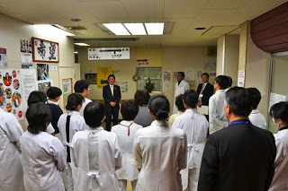 スーツ姿の佐藤町長が前で話しているのを、向かい合って立つ白衣を着た多くの医療スタッフが話を聞いている後ろ姿の写真