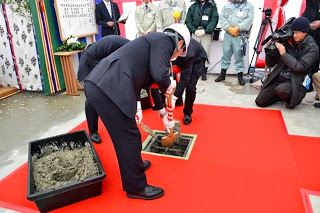 周囲には報道陣や関係者が見守り、赤いカーペットの上でスーツ姿の男性が儀式用のスコップで生コンクリートを穴に入れている様子が写っているコンクリート納入の儀の様子の写真