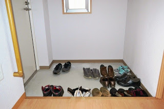 左側に手すりが設置された室内の玄関部分にさまざまな種類の靴がきれいに並べられている、左側に出入口の白いドアと奥に縦長の小窓が見える写真