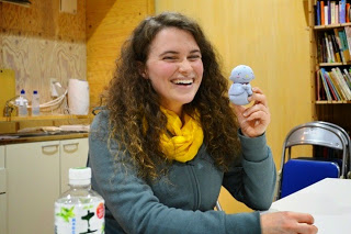 ペットボトルが置かれたテーブルの席に、左手にお地蔵様のぬいぐるみを持った黄色いマフラーを巻いた女性の先生が笑顔で座っている写真