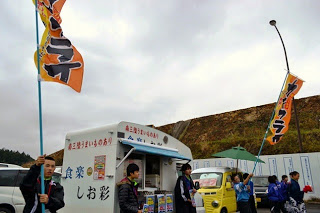 白いキッチンカーの前で「カキフライ」と書かれたオレンジ色の旗を立てながら中学生たちが呼び込みをしている様子の写真