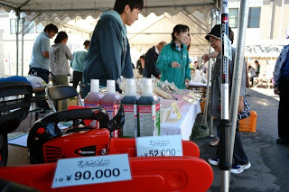 テントの下で、9万円や52,000円のポップがついたチェーンソーや農薬などの農業用品が並び、販売員が買い物客と会話している写真