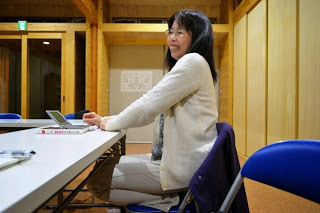 手元にはノートパソコンやノートが置かれている机の横に、白いカーディガンを着た女性が椅子に正座をして座って笑顔を見せている写真