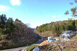 左に向かって続く緩やかなカーブの道路の両側に山林が広がり、右手前の造成地に建設中の住宅が見える写真