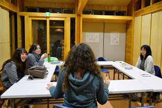 木の内装の部屋で4人の女性がロの字型に組まれた机を囲んで座り、手前に座っている先生が話ているのを他の女性たちが聞いている写真