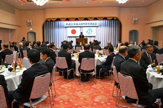 円卓が並ぶ広い宴会場で、多くのスーツ姿の男性たちが正面の壇上に注目しながら着席している写真