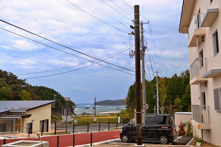 建物の間から海と空が見え、手前の駐車場には黒い車が駐車され、舗装された道路がある様子が写っている写真