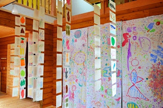 壁一面に子どもたちのカラフルな絵が描かれ、天井から野菜や果物などが描かれた沢山の絵が吊るされている写真