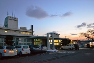 夕暮れ時の駐車場に車が並んでおり、建物に「志津川病院」と書かれた公立病院の全景が見える写真