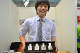 男性が白いシャツとネクタイ姿で笑顔を見せながら、四本の瓶詰め商品が並んだ黒いギフトボックスを両手で持って紹介している写真