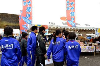 背中に「志津川」と大きく書かれた青いジャージ姿の中学生たちが、サケ祭りと書かれたのぼり旗が設置されたテントの前に集まっている様子の写真