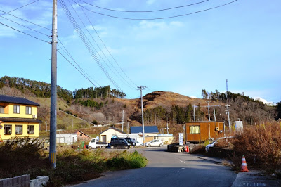 新しく整備された道路の周辺にプレハブ小屋や住宅が並び、背景に山が削られた造成地が見える街並みの写真