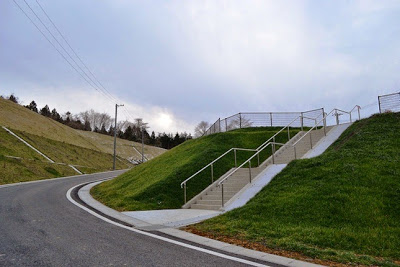 右に向かってカーブする緩やかな坂道沿いの緑の法面の斜面に、手すりつきのコンクリート製の階段が整備されている写真