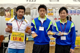 青と白のジャージを着た三人の男子生徒がテントの前で加工食品を手に持って笑顔で立っている写真