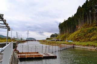 小高い山に囲まれた川に、金網とロープで区切られた人工のいけすのような設備が設置されている写真