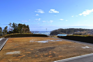 青空の下、海が望める高台にあり、周囲の道路や区画が整備された水たまりが点在している造成地の写真