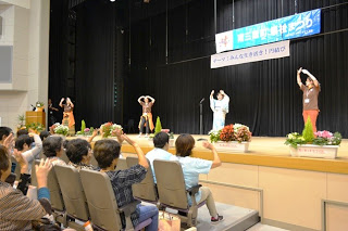 ステージ上で着物を着た女性を中央に、茶色のシャツを着た女性たちが手を挙げて体操をしており、観客席の人たちも一緒に同じ動きをしている写真