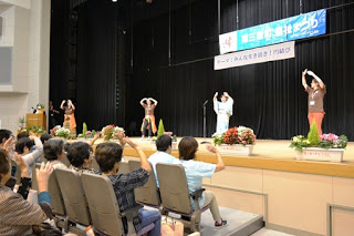 ホールのステージ上で4人が両手を上げて大きく丸をしている体操を披露し、観客席で見ている参加者も一緒に手を動かしている様子が写っている写真