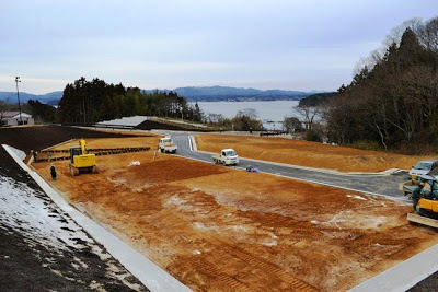 奥には海が広がっている高台に、整地された住宅区画で複数の建設車両が作業中の写真