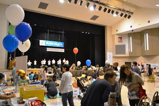 ステージではお揃いの白いシャツを着た人たちが並び、風船や商品が並ぶブースのある広い会場に多くの来場者が集まっている写真