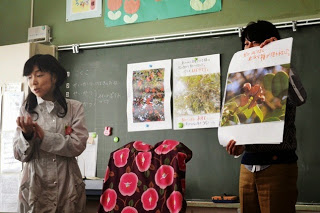 黒板の前で男性が木の実や植物の写真が印刷された大きなポスターを持って見せており、左側に立つ女性が子供たちに説明している授業の風景の写真