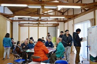 木の梁が見える室内で住民たちが椅子に座って話し合いを行っている地域住民の集まりの写真