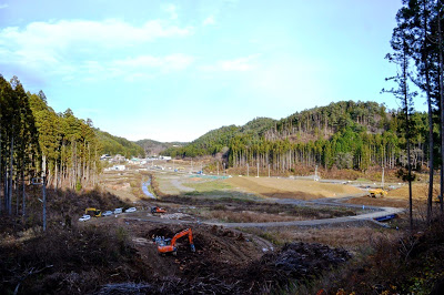 伐採された木々と造成中の土の斜面の間から谷あいに広がる復興工事現場を見渡した様子を高台から撮影した写真