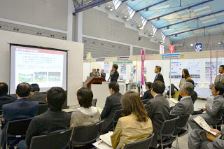 展示会場内の一角で、スーツ姿の発表者がスクリーンを使ってプレゼンテーションを行い、それを椅子に座った多くの聴衆が真剣に聞いている写真